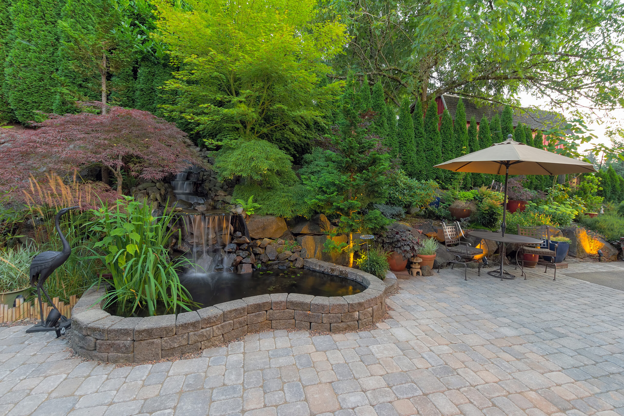 Serene backyard landscaping featuring a spacious patio surrounded by lush greenery and centered around a captivating waterfall pond. Serene backyard landscaping featuring a spacious patio surrounded by lush greenery and centered around a captivating waterfall pond.